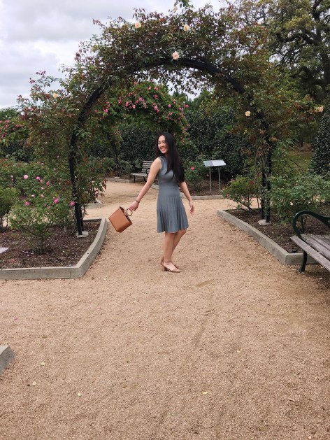 woman under rose arch wearing a sage dress while swinging a brown bag